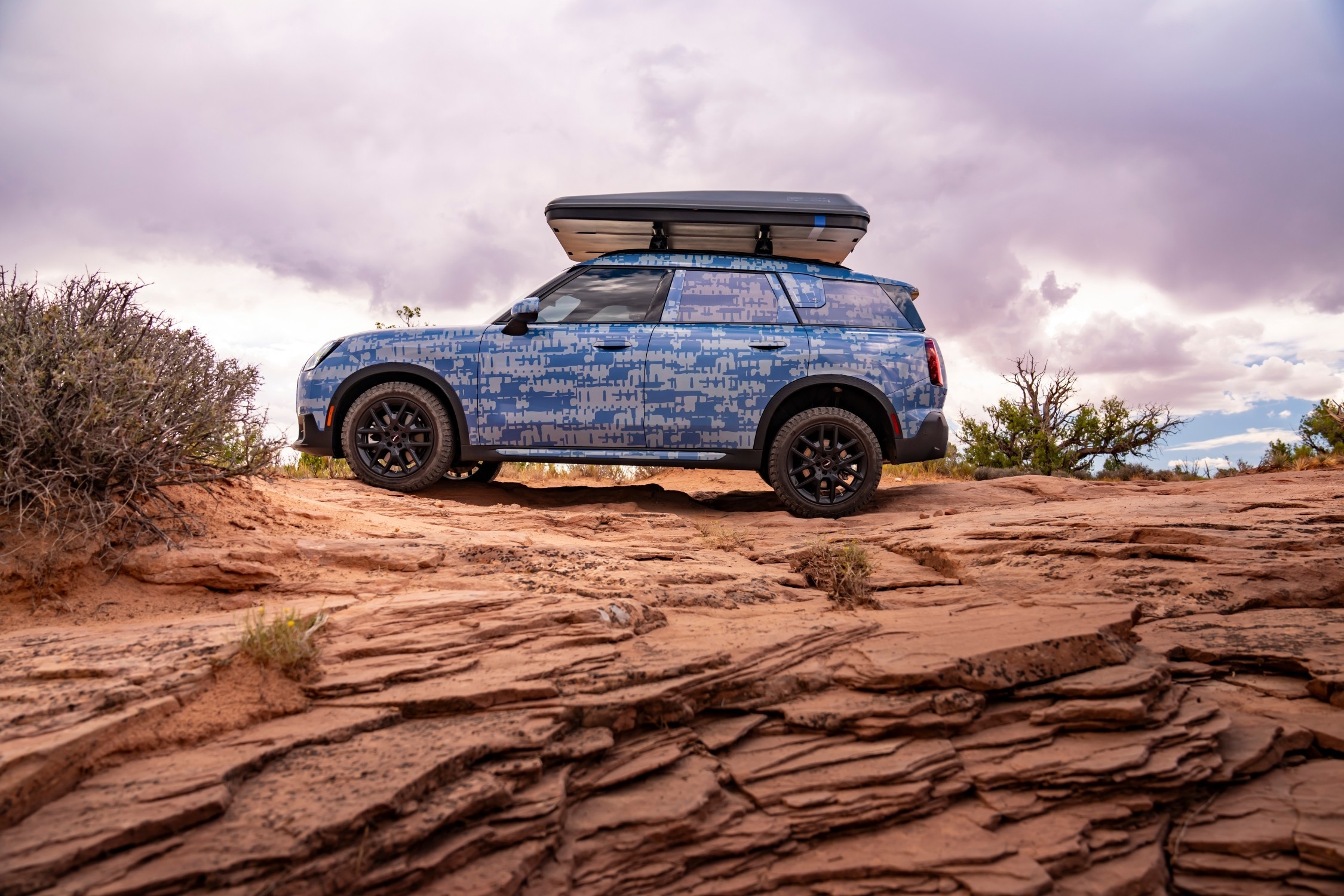 MINI Countryman visto de lado, com camuflagem e caixa de tejadilho, estacionado  sobre uma formação rochosa